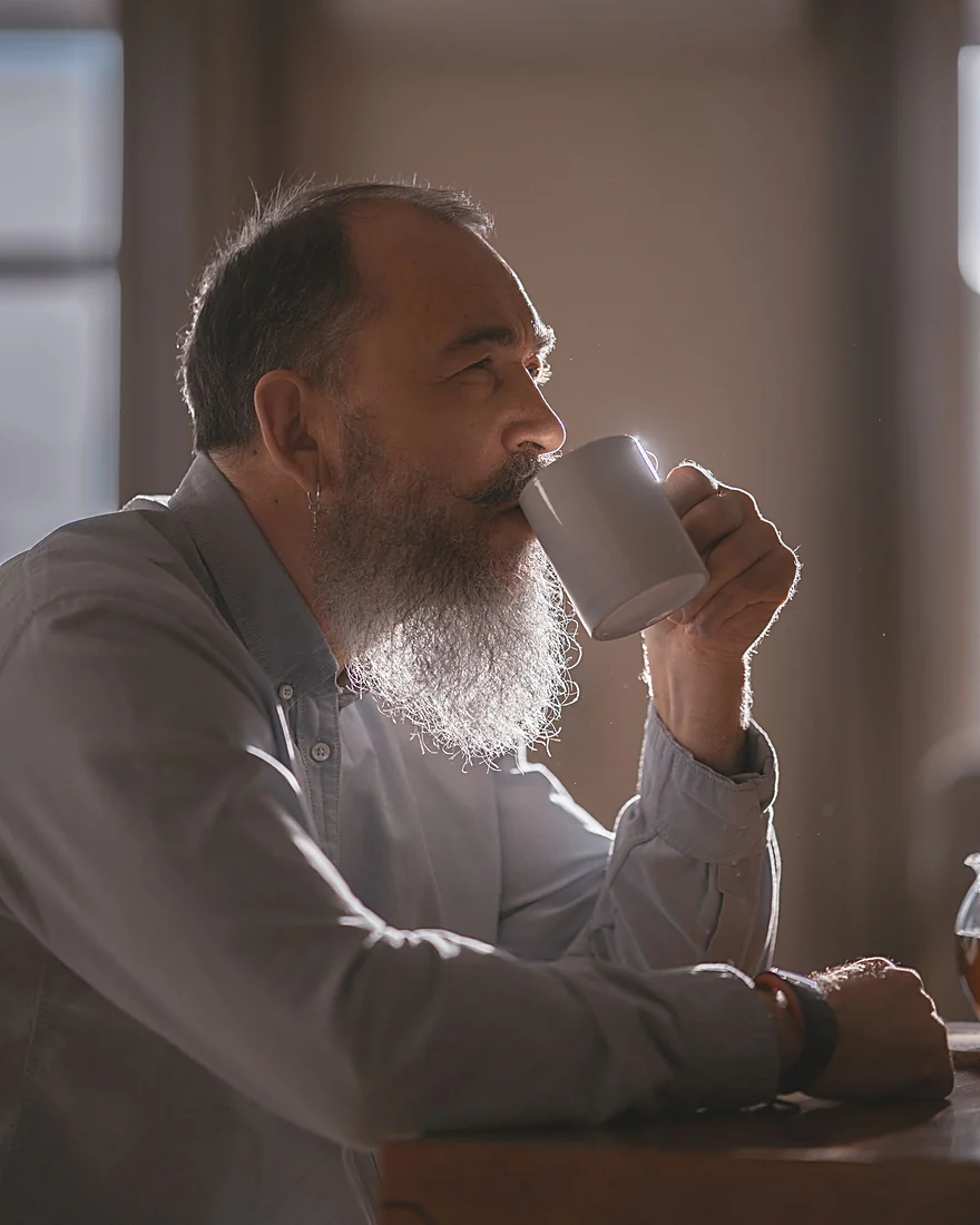 A bearded man sipping coffee from a white mug in warm morning light