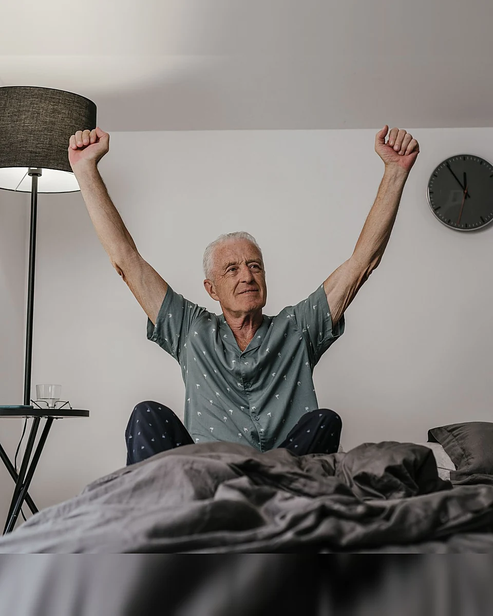 A man sitting up in bed at morning, arms stretched wide, having slept through the night