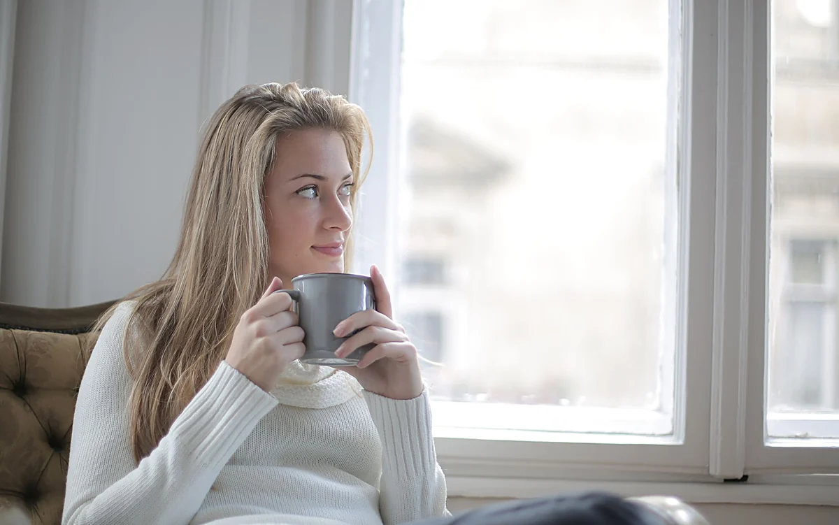 A woman by a bright window, holding a mug of coffee in both hands