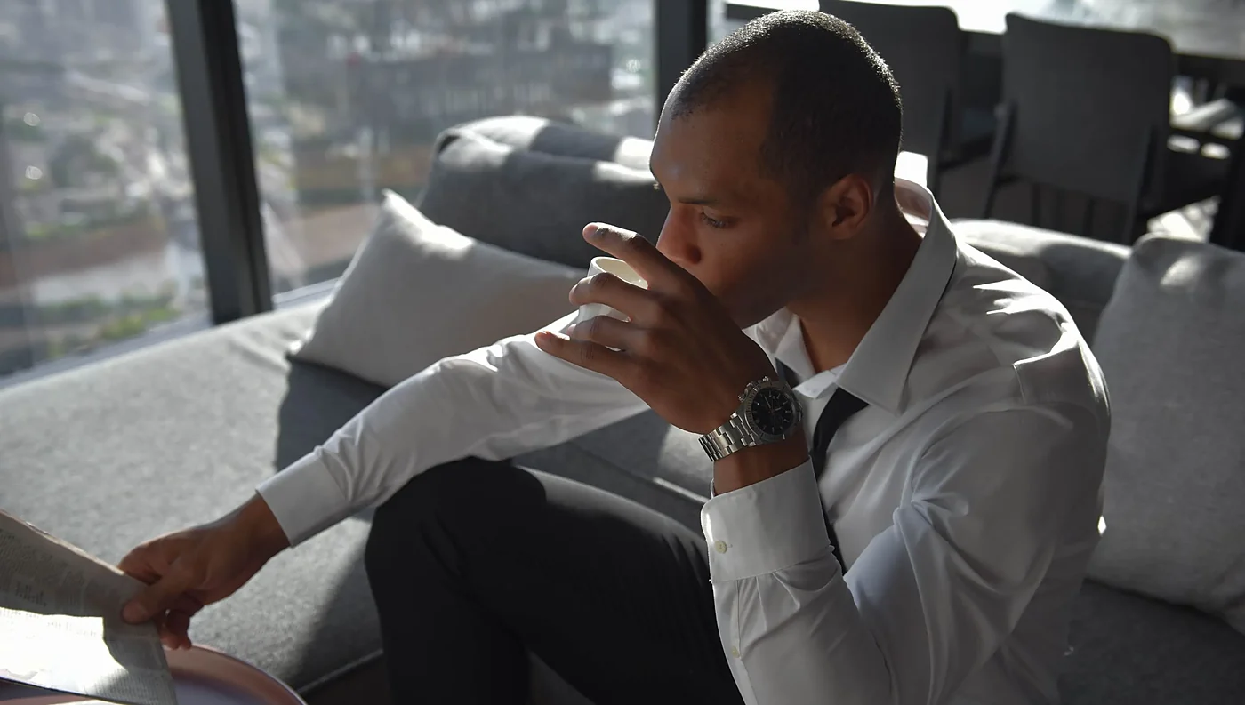 A man in a white shirt drinking coffee on a sofa by a large window