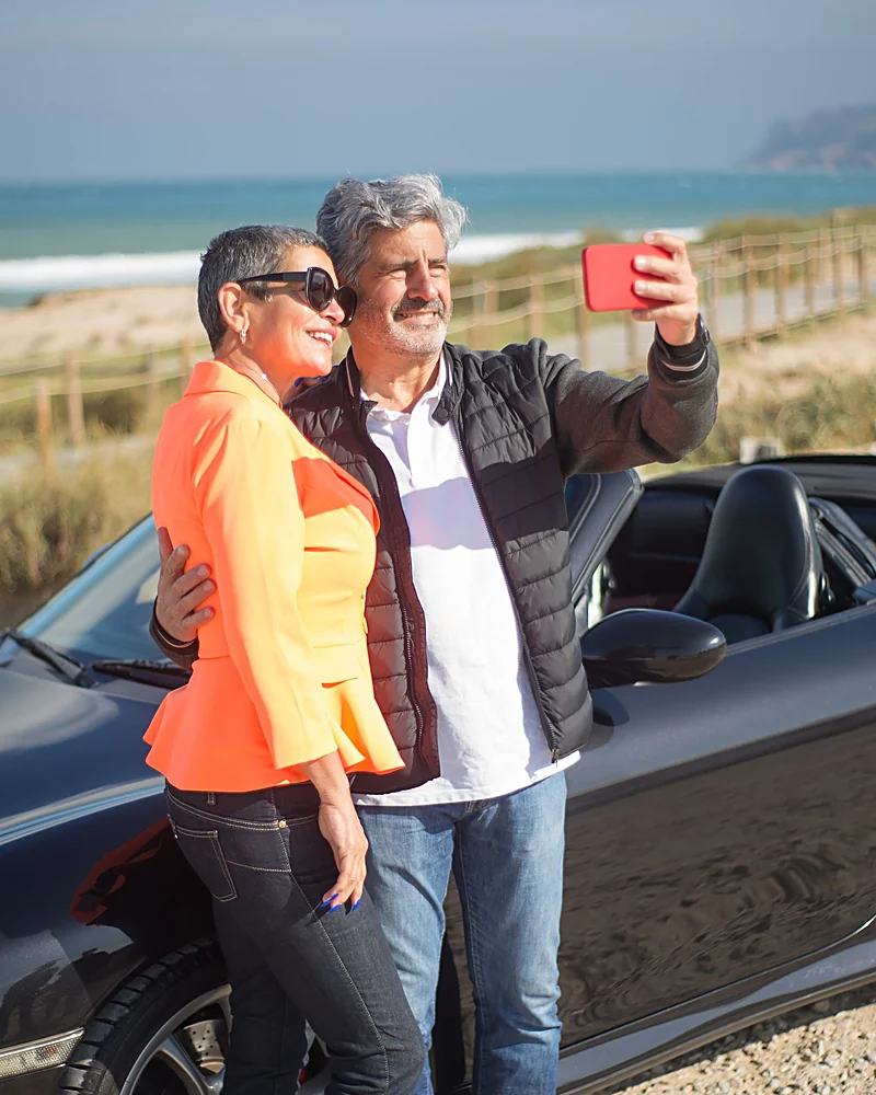 A couple in their sixties taking a selfie beside their car on a coastal road